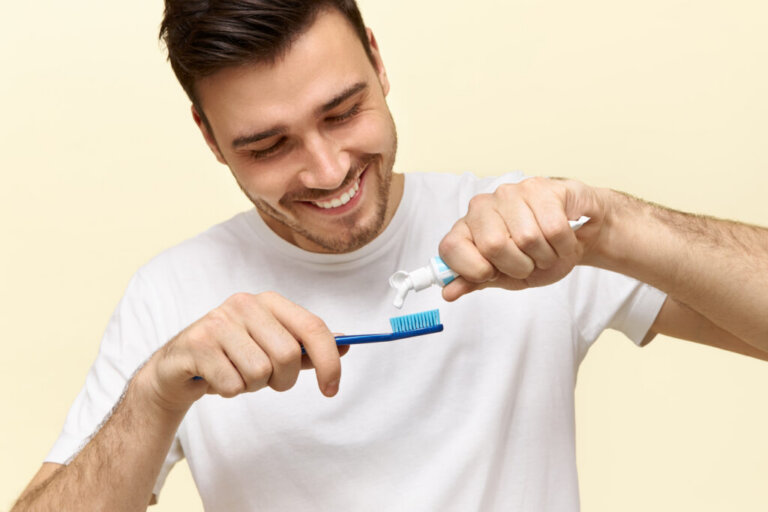 Close up of attractive young bearded man is brushing his teeth. He is standing and smiling. The man is squeezing tooth-paste into tooth-brush