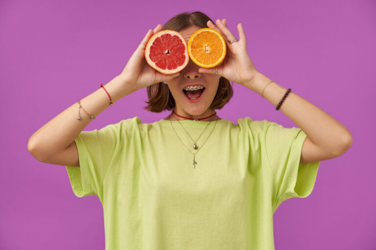 Female student, young lady with big smile, holding grapefruit and orange over her eyes. Standing over purple background